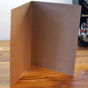 May include: A blank brown card with a folded edge. The card is sitting on a wooden surface.