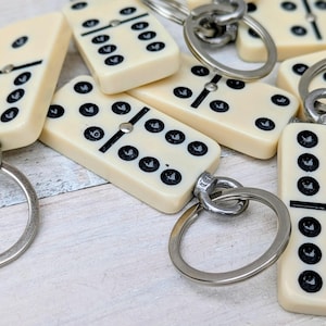 May include: Ivory domino keyrings with black dots and silver rings. The dominoes are rectangular and have a black line dividing the two sides. The keyrings are scattered on a light-coloured surface.