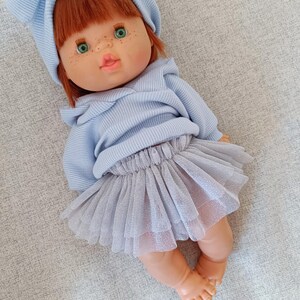 K&ouml;nnte beinhalten: Eine Puppe mit rotem Haar, gr&uuml;nen Augen und Sommersprossen, bekleidet mit einem hellblauen Kapuzenpullover, einem passenden Stirnband und einem grauen T&uuml;llrock. Die Puppe liegt auf einer grauen Oberfl&auml;che.