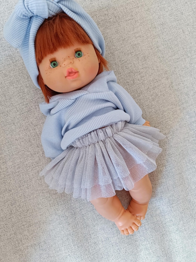 K&ouml;nnte beinhalten: Eine Puppe mit rotem Haar, gr&uuml;nen Augen und Sommersprossen, bekleidet mit einem hellblauen Kapuzenpullover, einem passenden Stirnband und einem grauen T&uuml;llrock. Die Puppe liegt auf einer grauen Oberfl&auml;che.