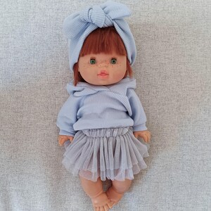 K&ouml;nnte beinhalten: Puppe mit rotem Haar, gr&uuml;nen Augen und Sommersprossen, bekleidet mit einem hellblauen Pullover, einem passenden Stirnband und einem grauen T&uuml;llrock. Die Puppe liegt auf einer grauen Oberfl&auml;che.