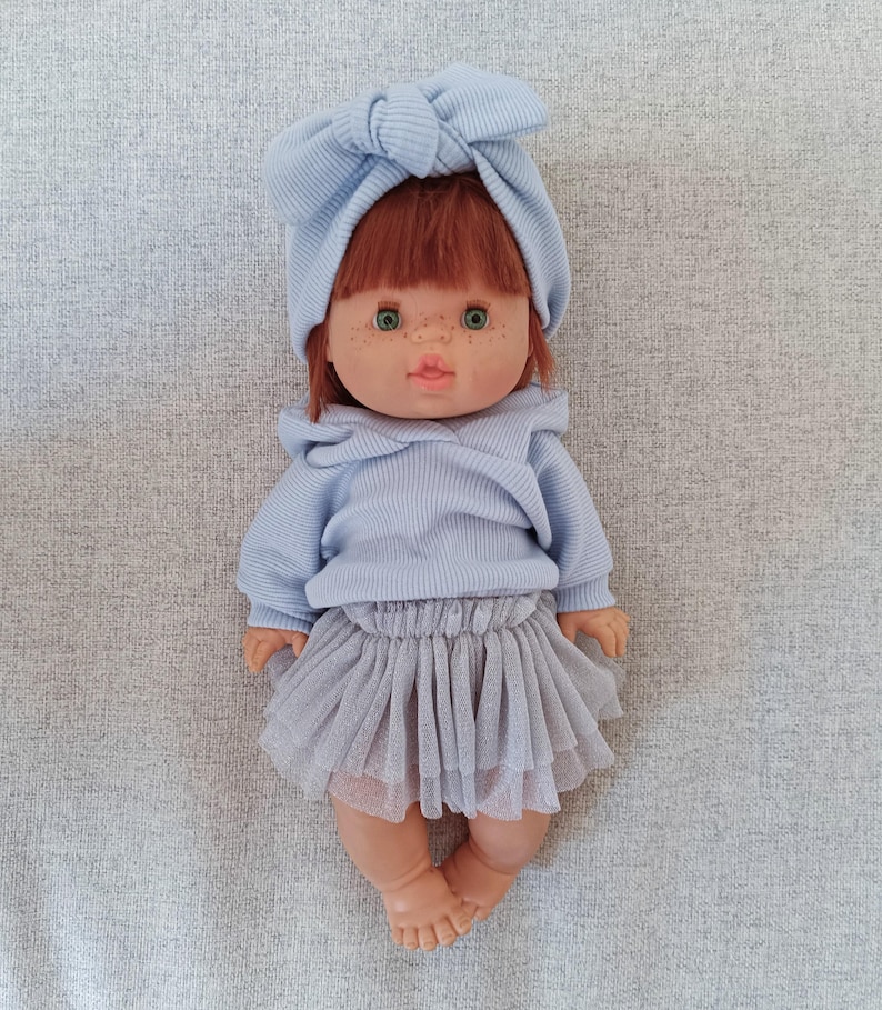 K&ouml;nnte beinhalten: Puppe mit rotem Haar, gr&uuml;nen Augen und Sommersprossen, bekleidet mit einem hellblauen Pullover, einem passenden Stirnband und einem grauen T&uuml;llrock. Die Puppe liegt auf einer grauen Oberfl&auml;che.