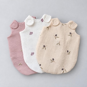 May include: Three sleeveless wearable blankets made of textured muslin fabric. The left one is dusty rose with small embroidered stars and moons. The middle one is white with purple floral sprigs. The right one is beige with olive branch prints.
