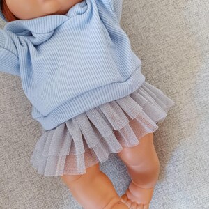 K&ouml;nnte beinhalten: Puppe mit roten Haaren und gr&uuml;nen Augen, bekleidet mit einem hellblauen Kapuzenpullover aus Rippstrick und einem grauen T&uuml;llrock. Die Beine und nackten F&uuml;&szlig;e der Puppe sind sichtbar. Die Puppe liegt auf einer grauen Oberfl&auml;che.