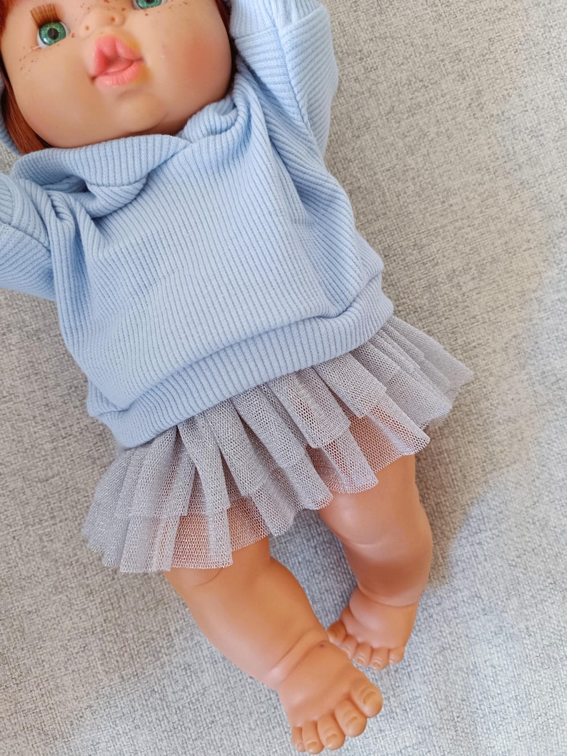 K&ouml;nnte beinhalten: Puppe mit roten Haaren und gr&uuml;nen Augen, bekleidet mit einem hellblauen Kapuzenpullover aus Rippstrick und einem grauen T&uuml;llrock. Die Beine und nackten F&uuml;&szlig;e der Puppe sind sichtbar. Die Puppe liegt auf einer grauen Oberfl&auml;che.