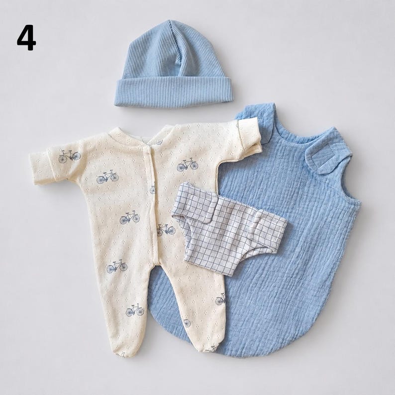 May include: A baby clothing set featuring a light blue ribbed beanie, a cream-colored long-sleeve footed sleeper with a blue bicycle print, a white and gray plaid diaper cover, and a sleeveless blue muslin swaddle sack with a side closure.