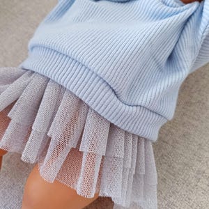 K&ouml;nnte beinhalten: Ein babyblaues Rippstrick-Oberteil mit einem mehrlagigen grauen T&uuml;llrock. Das Oberteil hat lange &Auml;rmel und einen Rundhalsausschnitt. Der Rock besteht aus mehreren Lagen weichem T&uuml;llstoff. Das Outfit ist f&uuml;r ein Baby oder Kleinkind konzipiert.