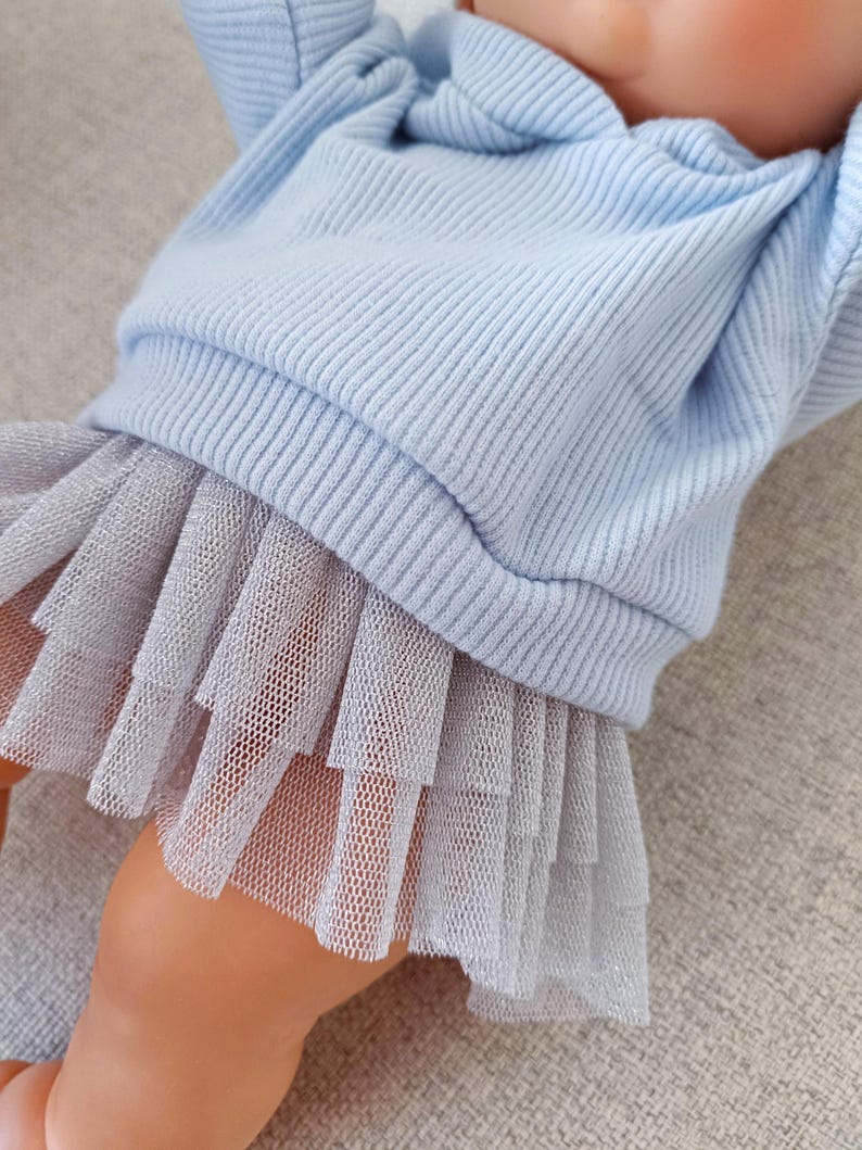 K&ouml;nnte beinhalten: Ein babyblaues Rippstrick-Oberteil mit einem mehrlagigen grauen T&uuml;llrock. Das Oberteil hat lange &Auml;rmel und einen Rundhalsausschnitt. Der Rock besteht aus mehreren Lagen weichem T&uuml;llstoff. Das Outfit ist f&uuml;r ein Baby oder Kleinkind konzipiert.