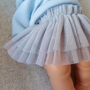 K&ouml;nnte beinhalten: Ein hellblauer Kapuzenpullover und ein mehrlagiger silberner T&uuml;llrock. Der Rock hat ein schimmerndes, glitzerndes Aussehen. Das Outfit ist f&uuml;r eine Puppe entworfen.