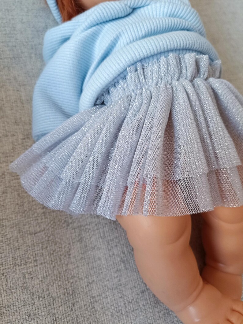 K&ouml;nnte beinhalten: Ein hellblauer Kapuzenpullover und ein mehrlagiger silberner T&uuml;llrock. Der Rock hat ein schimmerndes, glitzerndes Aussehen. Das Outfit ist f&uuml;r eine Puppe entworfen.