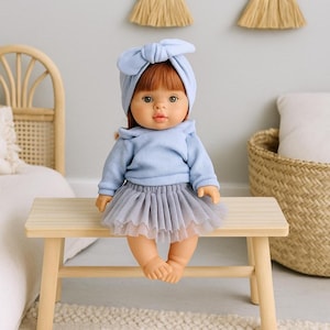 K&ouml;nnte beinhalten: Eine Puppe mit roten Haaren und gr&uuml;nen Augen, die einen hellblauen Pullover und ein passendes Stirnband tr&auml;gt. Die Puppe sitzt auf einer kleinen Holzbank und tr&auml;gt einen grauen T&uuml;llrock. Die Puppe ist ein Spielzeug.