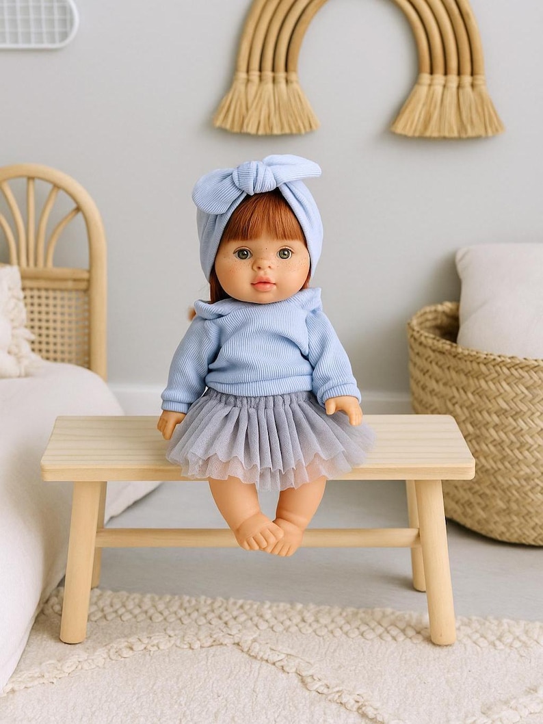 K&ouml;nnte beinhalten: Eine Puppe mit roten Haaren und gr&uuml;nen Augen, die einen hellblauen Pullover und ein passendes Stirnband tr&auml;gt. Die Puppe sitzt auf einer kleinen Holzbank und tr&auml;gt einen grauen T&uuml;llrock. Die Puppe ist ein Spielzeug.