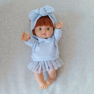 K&ouml;nnte beinhalten: Eine Puppe mit roten Haaren, gr&uuml;nen Augen und Sommersprossen, bekleidet mit einem hellblauen Outfit. Das Outfit umfasst einen Pullover, einen T&uuml;llrock und ein passendes Schleifen-Stirnband. Die Puppe liegt mit erhobenen Armen.