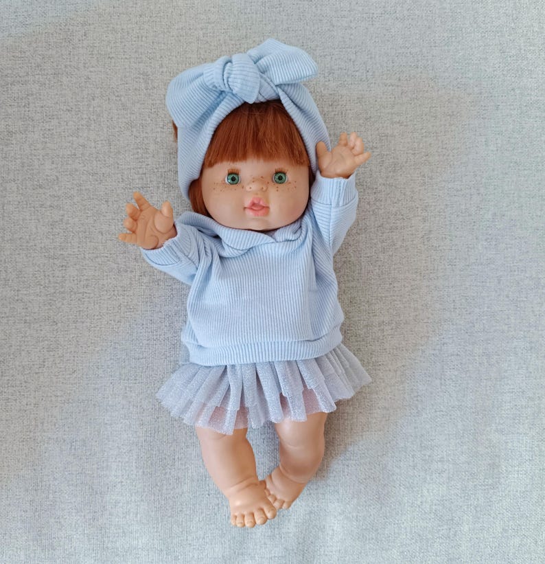 K&ouml;nnte beinhalten: Eine Puppe mit roten Haaren, gr&uuml;nen Augen und Sommersprossen, bekleidet mit einem hellblauen Outfit. Das Outfit umfasst einen Pullover, einen T&uuml;llrock und ein passendes Schleifen-Stirnband. Die Puppe liegt mit erhobenen Armen.