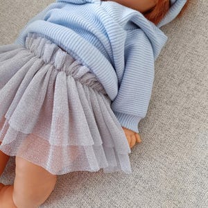 K&ouml;nnte beinhalten: Eine Puppe mit einem hellblauen Kapuzenpullover und einem mehrlagigen grauen T&uuml;llrock. Die Puppe hat rotes Haar und liegt auf einer grauen Oberfl&auml;che. Das Outfit ist aus einem weichen, bequemen Material.