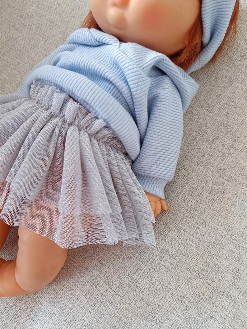 K&ouml;nnte beinhalten: Eine Puppe mit einem hellblauen Kapuzenpullover und einem mehrlagigen grauen T&uuml;llrock. Die Puppe hat rotes Haar und liegt auf einer grauen Oberfl&auml;che. Das Outfit ist aus einem weichen, bequemen Material.