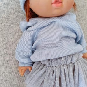 K&ouml;nnte beinhalten: Puppe mit rotem Haar, gr&uuml;nen Augen und Sommersprossen, bekleidet mit einem hellblauen Kapuzenpullover, einer passenden M&uuml;tze und einem silbernen T&uuml;llrock. Das Outfit der Puppe hat ein weiches Pastellfarbschema.