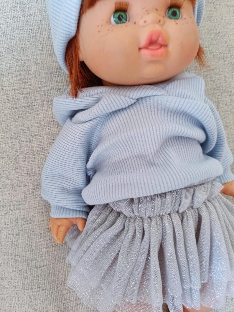 K&ouml;nnte beinhalten: Puppe mit rotem Haar, gr&uuml;nen Augen und Sommersprossen, bekleidet mit einem hellblauen Kapuzenpullover, einer passenden M&uuml;tze und einem silbernen T&uuml;llrock. Das Outfit der Puppe hat ein weiches Pastellfarbschema.