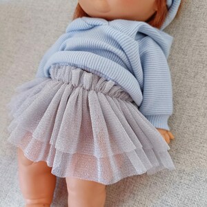 K&ouml;nnte beinhalten: Eine Puppe mit hellblauem Kapuzenpullover und einem mehrlagigen, silbergrauen T&uuml;llrock. Die Puppe hat rotes Haar, gr&uuml;ne Augen und ein passendes blaues Stirnband. Die Puppe liegt auf einer grauen Oberfl&auml;che.