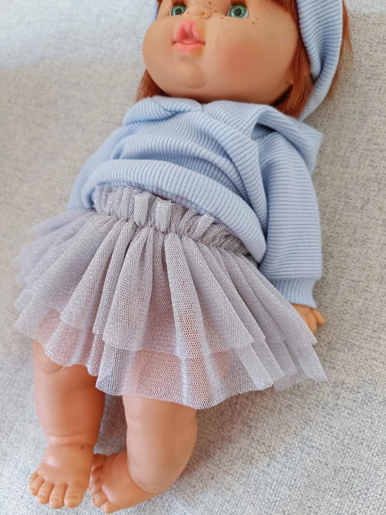 K&ouml;nnte beinhalten: Eine Puppe mit hellblauem Kapuzenpullover und einem mehrlagigen, silbergrauen T&uuml;llrock. Die Puppe hat rotes Haar, gr&uuml;ne Augen und ein passendes blaues Stirnband. Die Puppe liegt auf einer grauen Oberfl&auml;che.