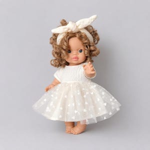 Peut inclure: Une poupée aux cheveux bruns bouclés portant un bandeau crème avec un nœud. La poupée est vêtue d'un haut à manches courtes de couleur crème et d'une jupe en tulle superposée avec un motif répétitif de petits cœurs blancs sur le tissu.