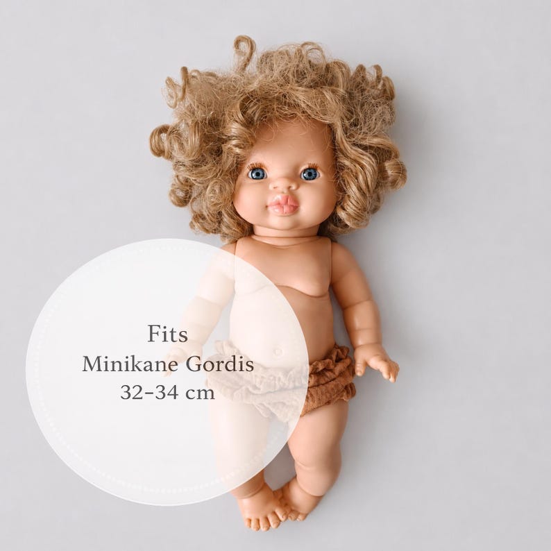 May include: A doll with curly light brown hair, blue eyes, and pink lips, wearing brown ruffled bloomers. A circular overlay on the image contains the text: Fits Minikane Gordis 32-34 cm. The doll has a light skin tone and a neutral facial expression.