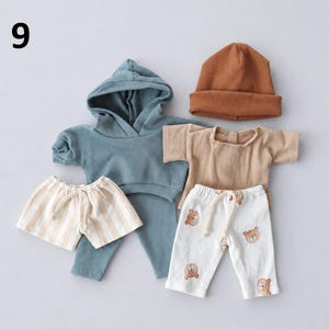 May include: A set of baby clothing including a blue ribbed hooded long-sleeve top, a blue ribbed pair of pants, a tan ribbed beanie, a tan short-sleeve shirt, tan and white striped shorts, and white pants with a brown bear face pattern.