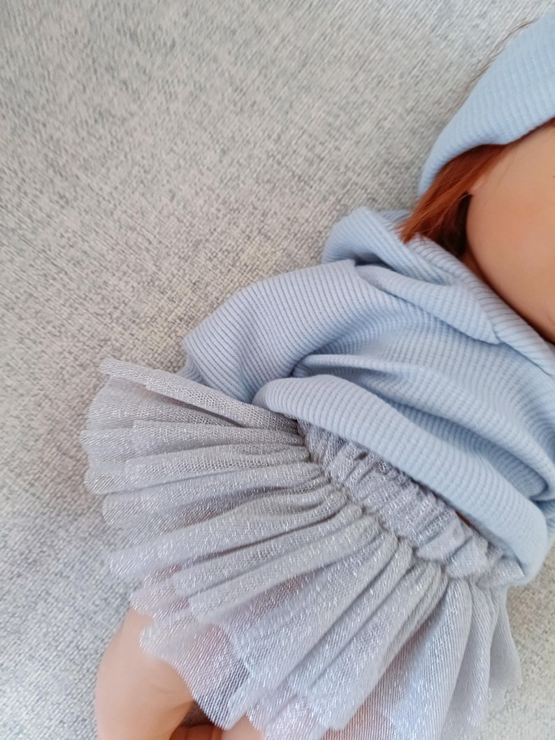 K&ouml;nnte beinhalten: Eine Puppe mit hellblauem Kapuzenpullover und passender M&uuml;tze. Der Arm der Puppe ist mit einem silbernen, mehrlagigen T&uuml;llrock geschm&uuml;ckt. Die Puppe hat rotes Haar und einen hellen Teint. Das Outfit ist weich und bequem.