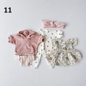May include: A set of baby clothing including a dusty pink short-sleeve hooded sweatshirt, a pair of striped bloomers, a white footed sleeper with brown hearts, a floral print dress with green and pink flowers, and a matching dusty pink headband.