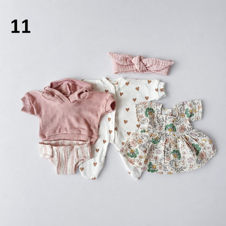 May include: A set of baby clothing including a dusty pink short-sleeve hooded sweatshirt, a pair of striped bloomers, a white footed sleeper with brown hearts, a floral print dress with green and pink flowers, and a matching dusty pink headband.