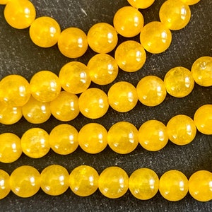 May include: A strand of bright yellow round beads. The beads are smooth and glossy, with a slight translucence. The beads are arranged in a staggered pattern, creating a visually appealing texture.