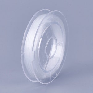 May include: A spool of clear elastic thread wound around a clear plastic bobbin. The thread is white and appears to be very thin.