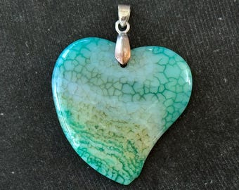Puede incluir: Colgante en forma de corazón con una piedra de ágata turquesa y verde amarillento. La piedra tiene un patrón agrietado único y está unida a una fianza plateada. El colgante es una joya.