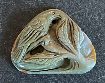 May include: A carved stone pendant featuring a bird perched on a branch with leaves. The pendant is made of a light green and brown stone with a smooth, polished finish.