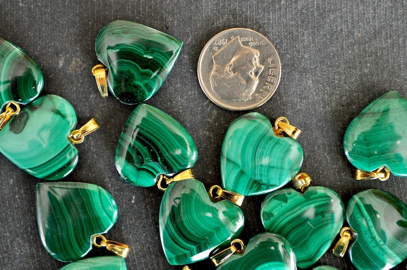 May include: A group of green malachite heart-shaped pendants with gold jump rings. The pendants are arranged on a dark gray surface. A US quarter is included for size reference.