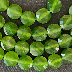 May include: Close-up of a strand of faceted, round, olive-green gemstone beads. The beads are strung together with green thread, and each bead has a geometric, multi-faceted surface. The beads are a vibrant, translucent green color.