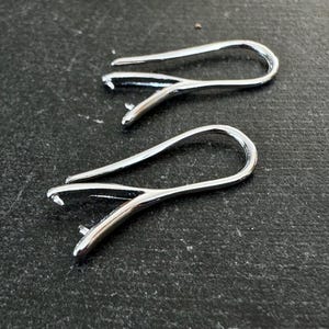 May include: Two silver-toned earring hooks, also known as ear wires, are shown on a dark grey surface. The hooks are made of metal and have a smooth, polished finish.