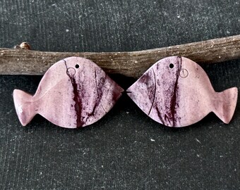 May include: Two pink and purple fish-shaped stone ornaments with black markings. The ornaments are facing each other with their mouths touching.