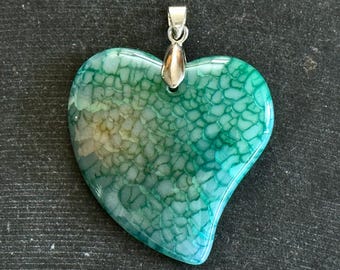 Puede incluir: Un colgante de corazón de ágata verde y blanca con una anilla de plata. La ágata tiene un patrón único y arremolinado.