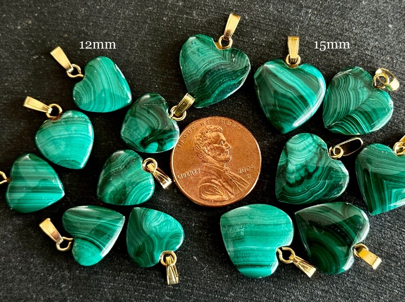 May include: A collection of 12mm and 15mm green malachite heart-shaped pendants with gold-tone bail. The pendants are arranged on a dark gray surface with a penny for scale.