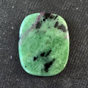 May include: A polished, oval-shaped gemstone pendant. The stone is primarily green with black and maroon inclusions. A small hole is drilled at the top for stringing. The pendant is set against a dark, textured background.