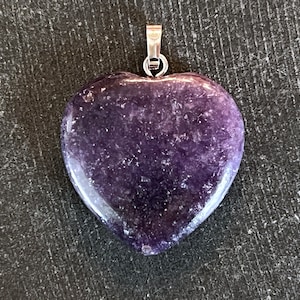 May include: A heart-shaped pendant made of a deep purple stone. The pendant has a polished surface and a silver-colored bail for attaching to a chain. The stone has a speckled appearance.