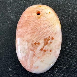 以下が含まれることがあります： 穴の開いた楕円形の宝石ペンダント。 桃色、クリーム色、オレンジ色の色合いで、独特のテクスチャパターンが特徴です。 小さな正方形のオレンジ色のインクルージョンが表面に散らばっています。 ペンダントの長さは約5cmです。