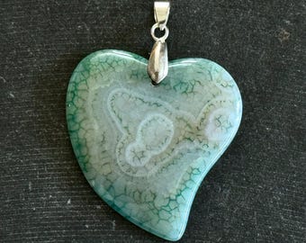 Puede incluir: Un colgante de corazón de ágata verde y blanca con una anilla de plata. La ágata tiene un patrón de remolino de verde y blanco.