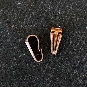 Op de afbeelding: Twee kleine, roségoudkleurige sieradssluitingen op een donker, getextureerd oppervlak. Eén sluiting heeft een gebogen haakvorm en de andere een rechthoekig ontwerp met verticale groeven. De sluitingen zijn waarschijnlijk voor kettingen of armbanden.