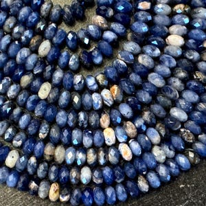 May include: Close-up of a collection of faceted, oval-shaped beads in shades of blue, with some beads appearing white or tan. The beads are strung together, creating a textured surface. The beads are likely for jewelry making.