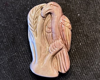 May include: A carved, off-white and pink eagle figurine. The eagle is perched on a tree branch, with detailed feather and wing carvings. The figurine has a smooth, polished finish and is likely made of ceramic or a similar material.
