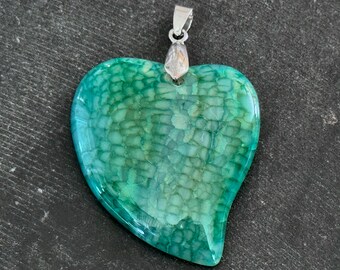 Puede incluir: Un colgante de corazón de ágata verde con una anilla de plata. La ágata tiene un patrón de remolino que se asemeja a la piel de cocodrilo.