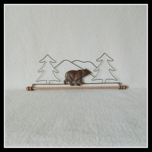 May include: A brown bear walking through a wire sculpture of a forest scene with two trees and mountains. The sculpture is mounted on a wooden dowel rod.