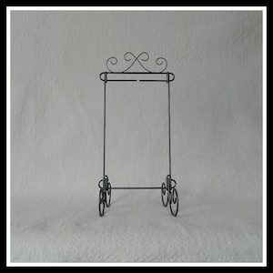 May include: Black metal easel stand with a decorative scroll design at the top and bottom. The stand is designed to hold a picture or other artwork.